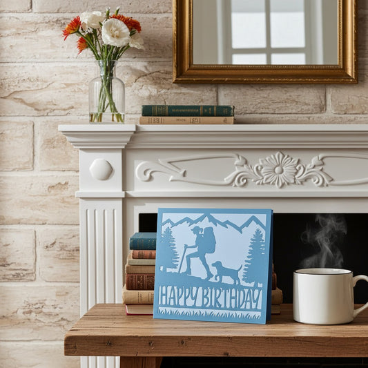 A square blue and white 'HAPPY BIRTHDAY' card featuring a silhouette of a hiker and dog, styled on a wooden table next to a white mug in front of a white fireplace mantel with a brick wall background.