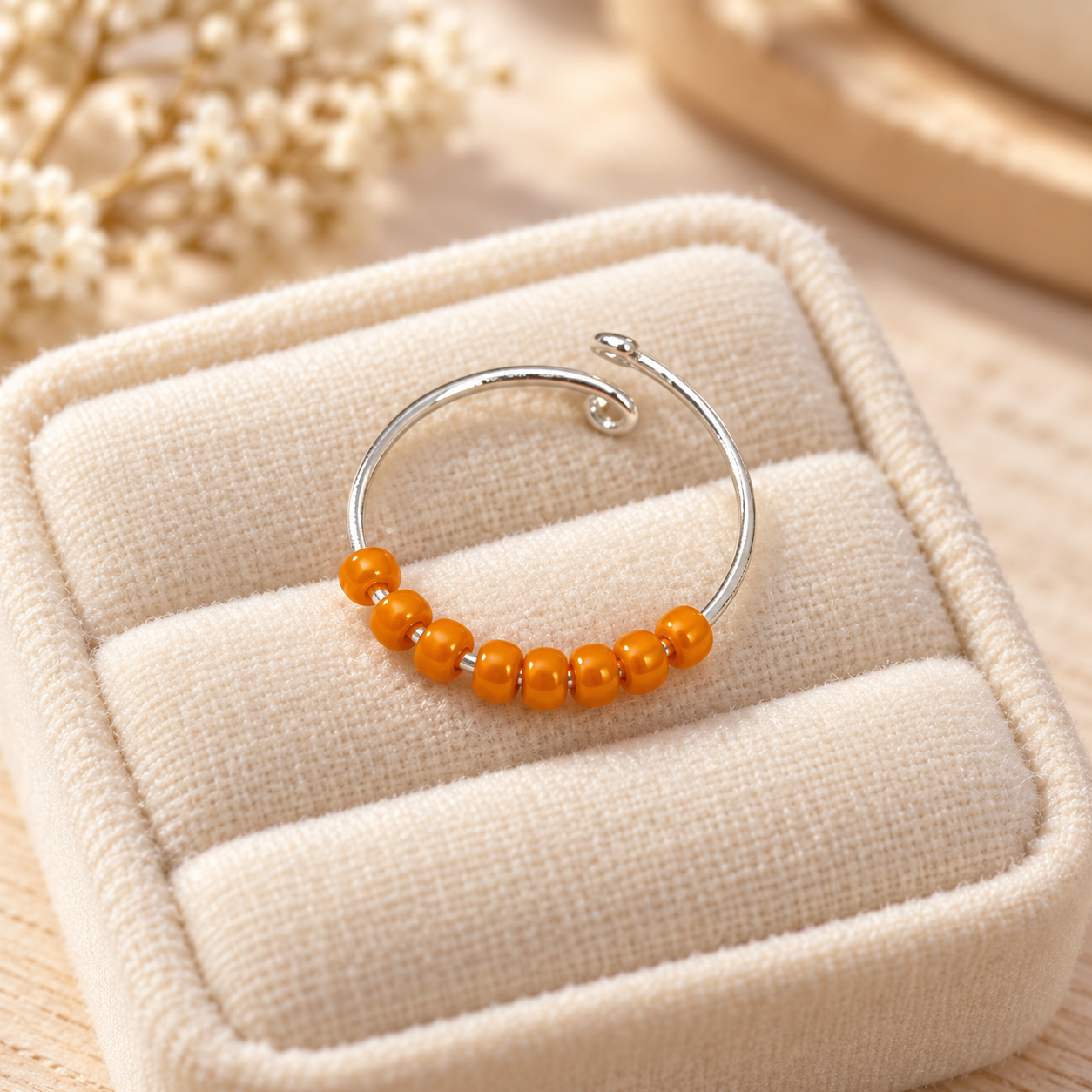 A silver-toned fidget ring with a row of small orange beads, displayed on a cream-coloured textured jewellery pouch.