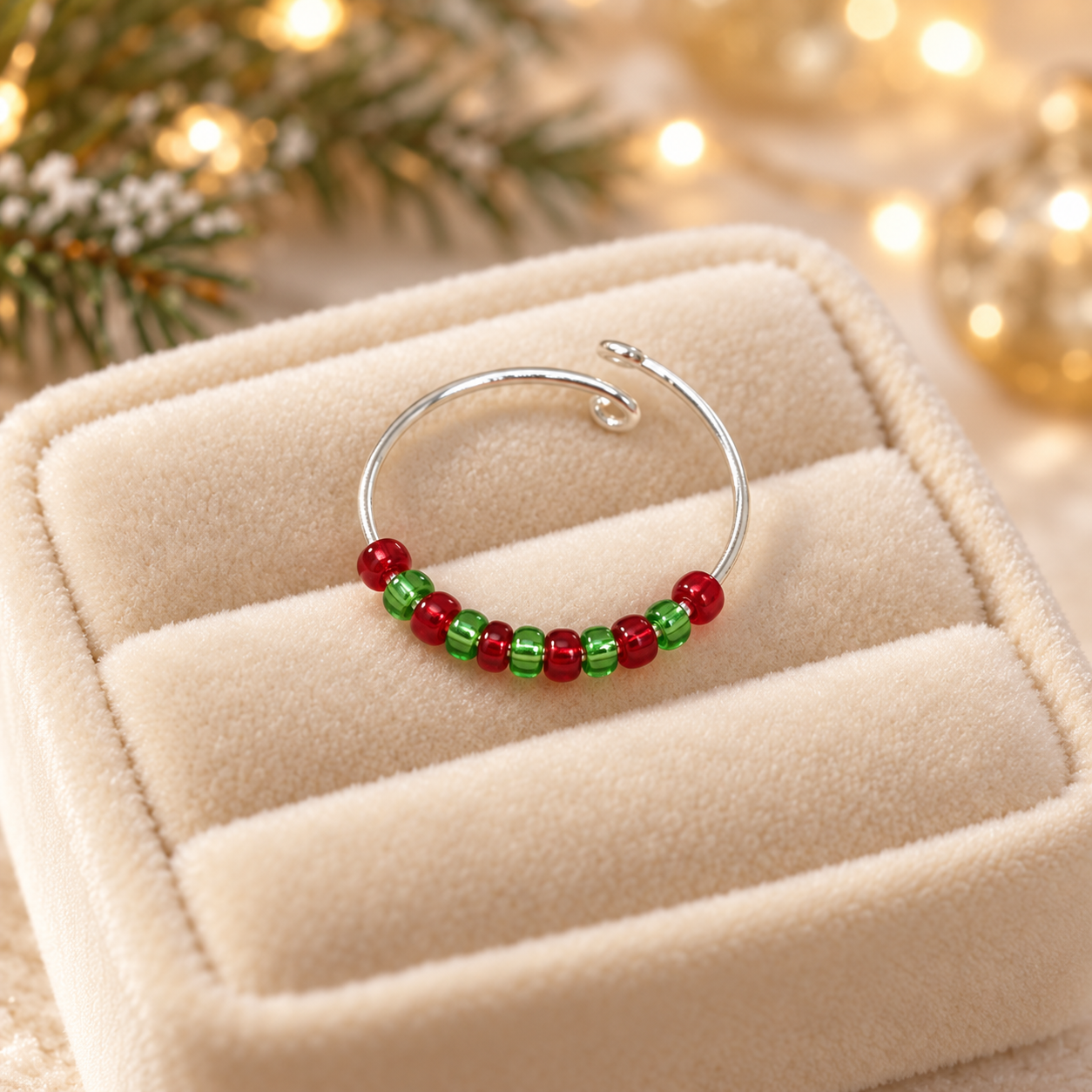 A silver-toned fidget ring with small red and green beads, displayed on a cream-coloured velvet jewellery box with a warm, festive bokeh background.