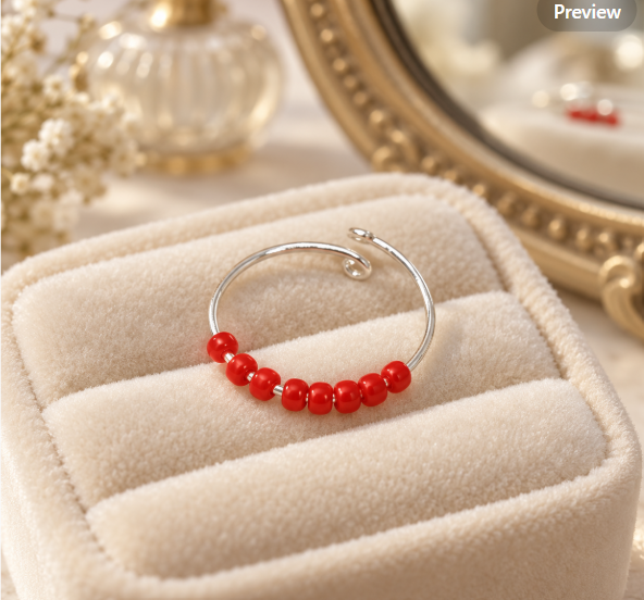 A silver-toned open fidget ring with a row of small red beads, displayed on a cream velvet jewellery box. The background features a gold mirror and soft decorative elements.