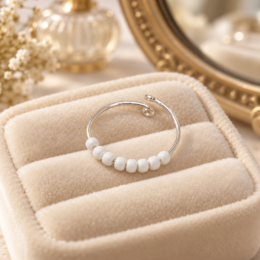 A silver-toned open fidget ring with a row of small white beads, displayed on a beige velvet jewellery box.