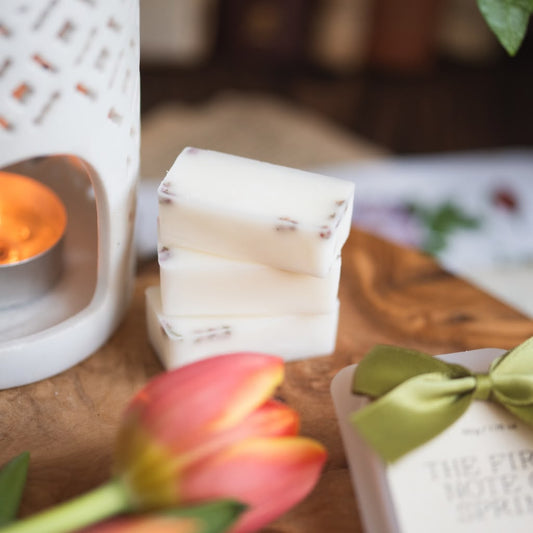 Three stacked white rectangular bars of wax melts with small dark inclusions, displayed on a wooden surface next to a white candle holder with a lit tea light, pink tulips, and a gift box with a green ribbon.