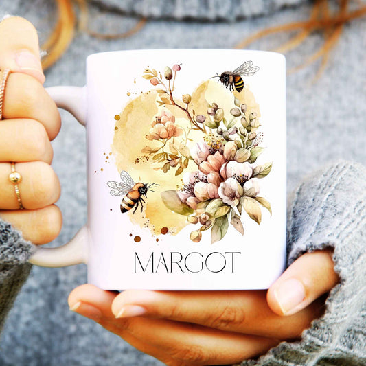 Person holding a bee mug with floral design and 'MARGOT' text