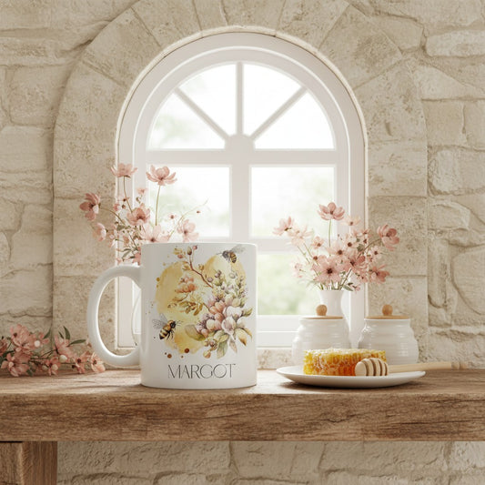 Mug with bee floral design and 'MARGOT' text on a wooden surface with flowers and a window in the background.