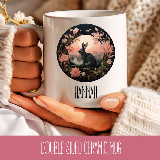 A ceramic mug with a floral design featuring a rabbit, held in a hand with a knitted sweater and a floral background.