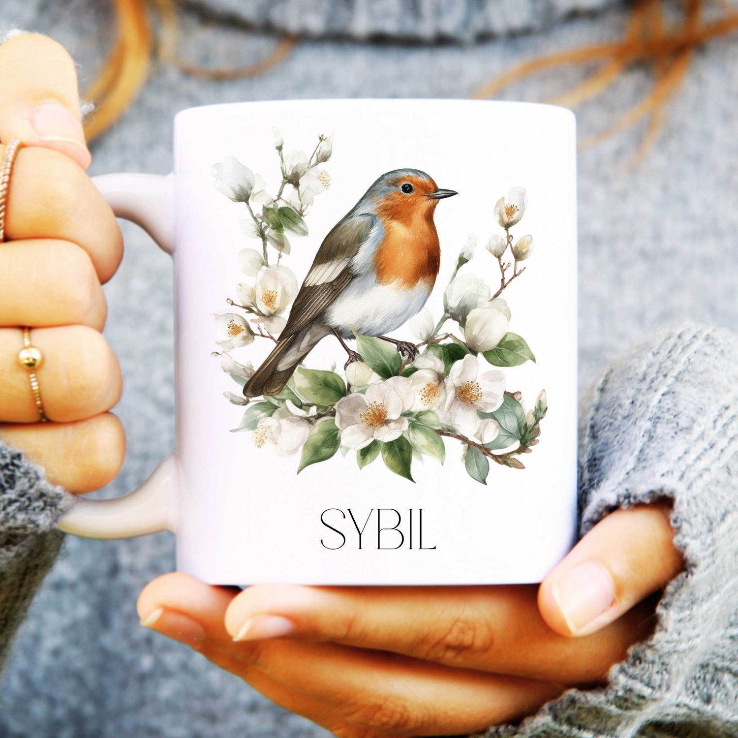 Mug with a robin and floral design held by a person wearing a grey sweater.