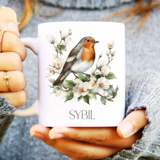 Mug with a robin and floral design held by a person wearing a grey sweater.