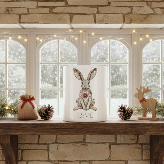 Decorative mug with floral hare design on a wooden table in front of a stone wall and window with Christmas decorations.