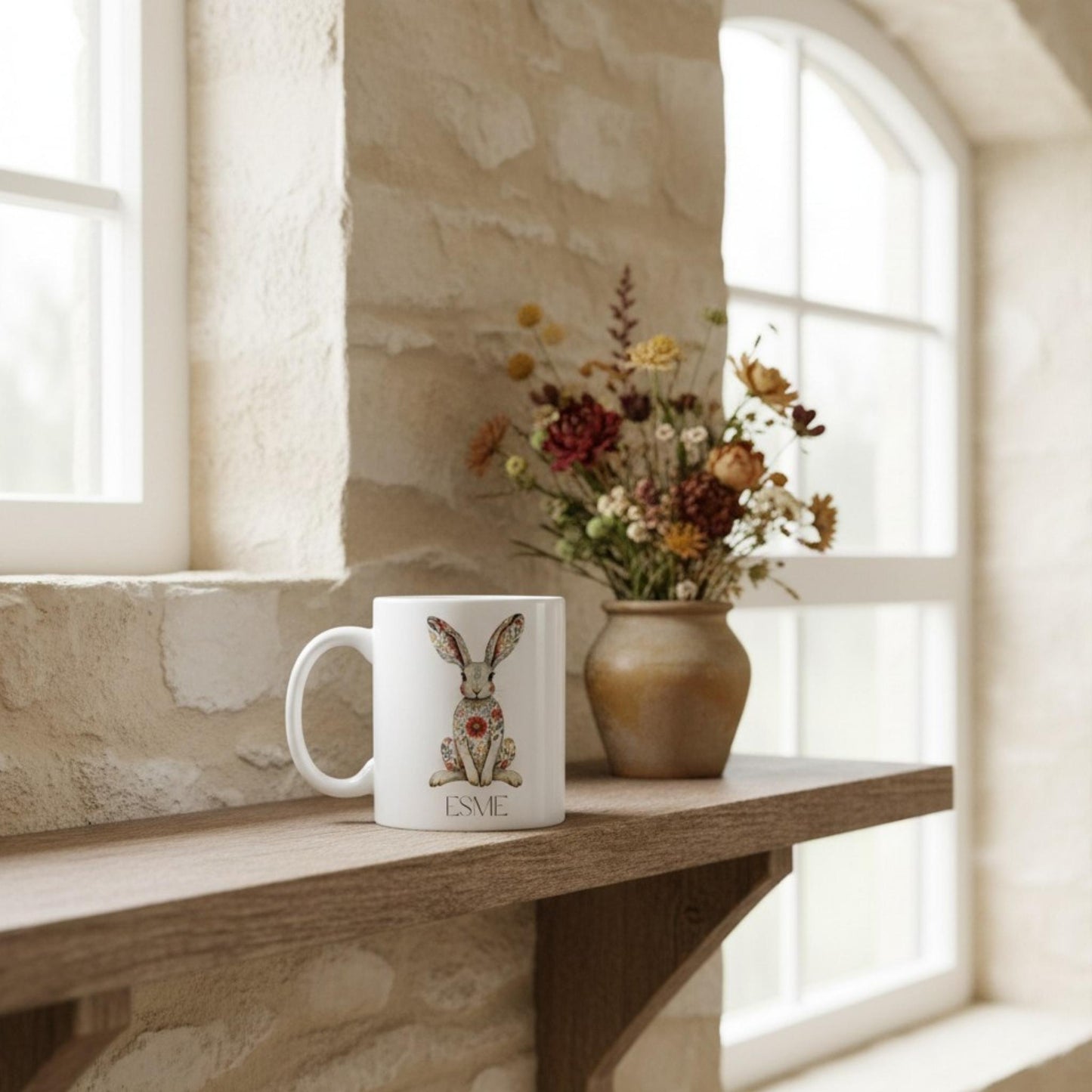 White mug with hare design and 'Esme' on a wooden shelf with flowers in the background.
