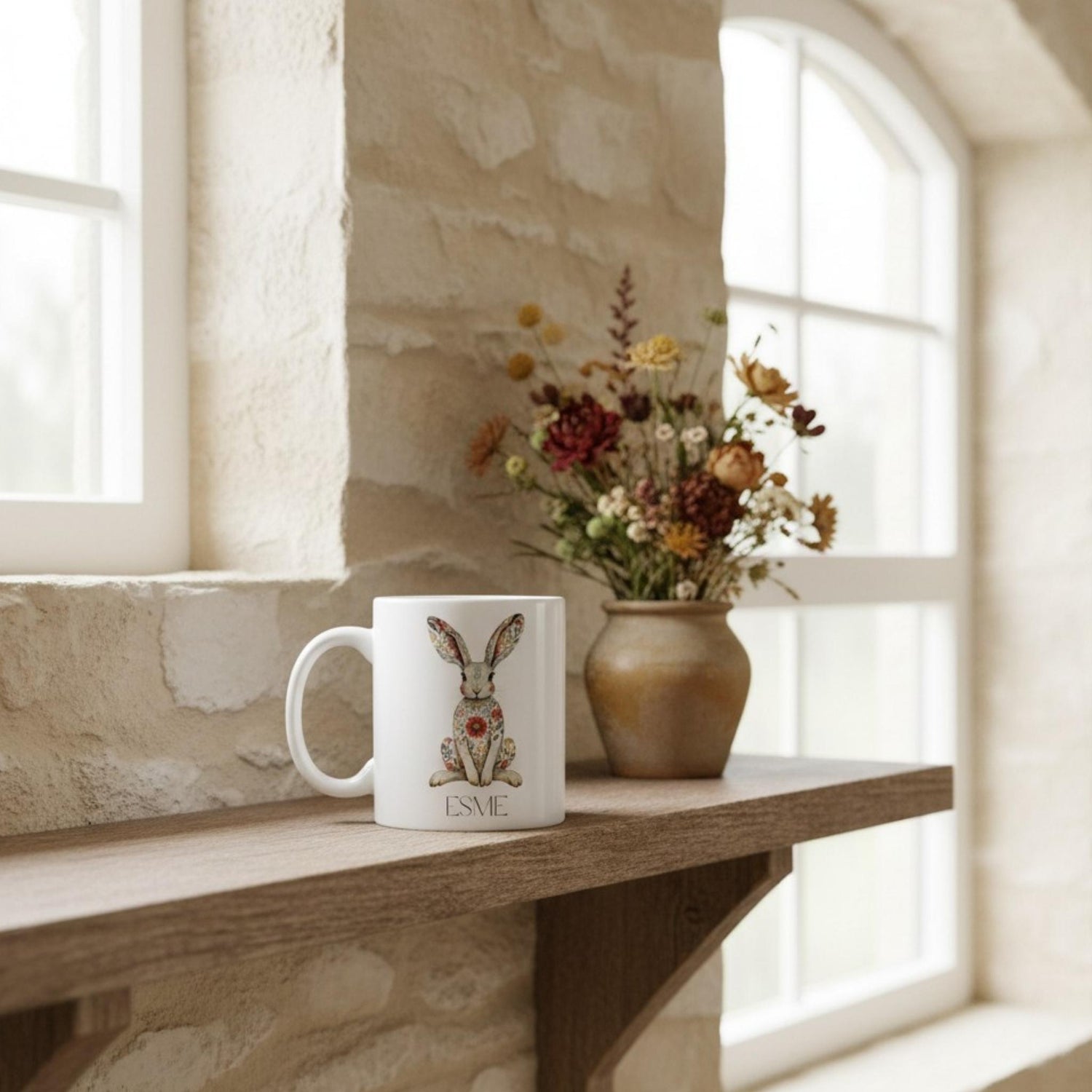 White mug with hare design and 'Esme' on a wooden shelf with flowers in the background.