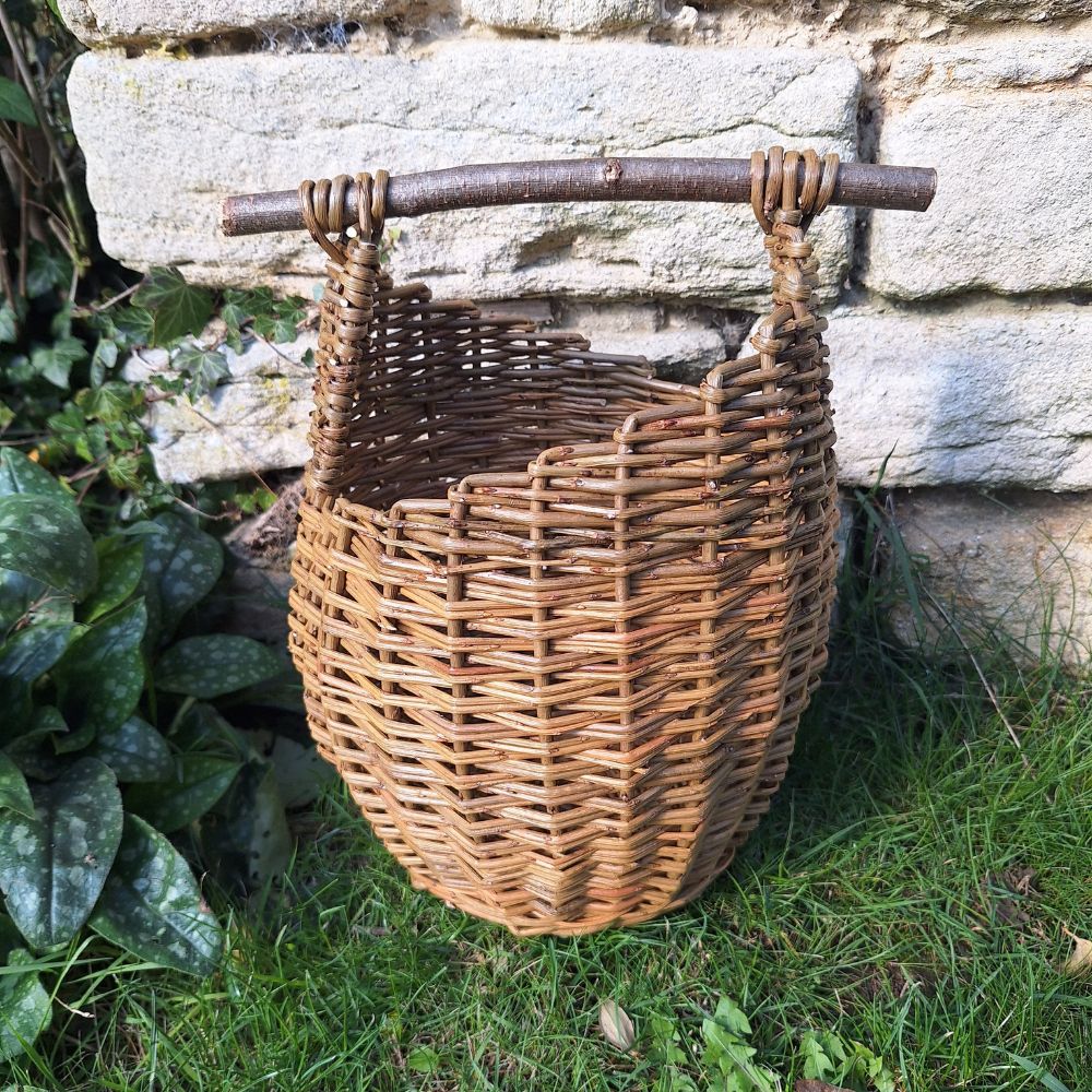 A handmade willow asymmetric top basket with a branch handle, set against a stone wall background with green foliage.