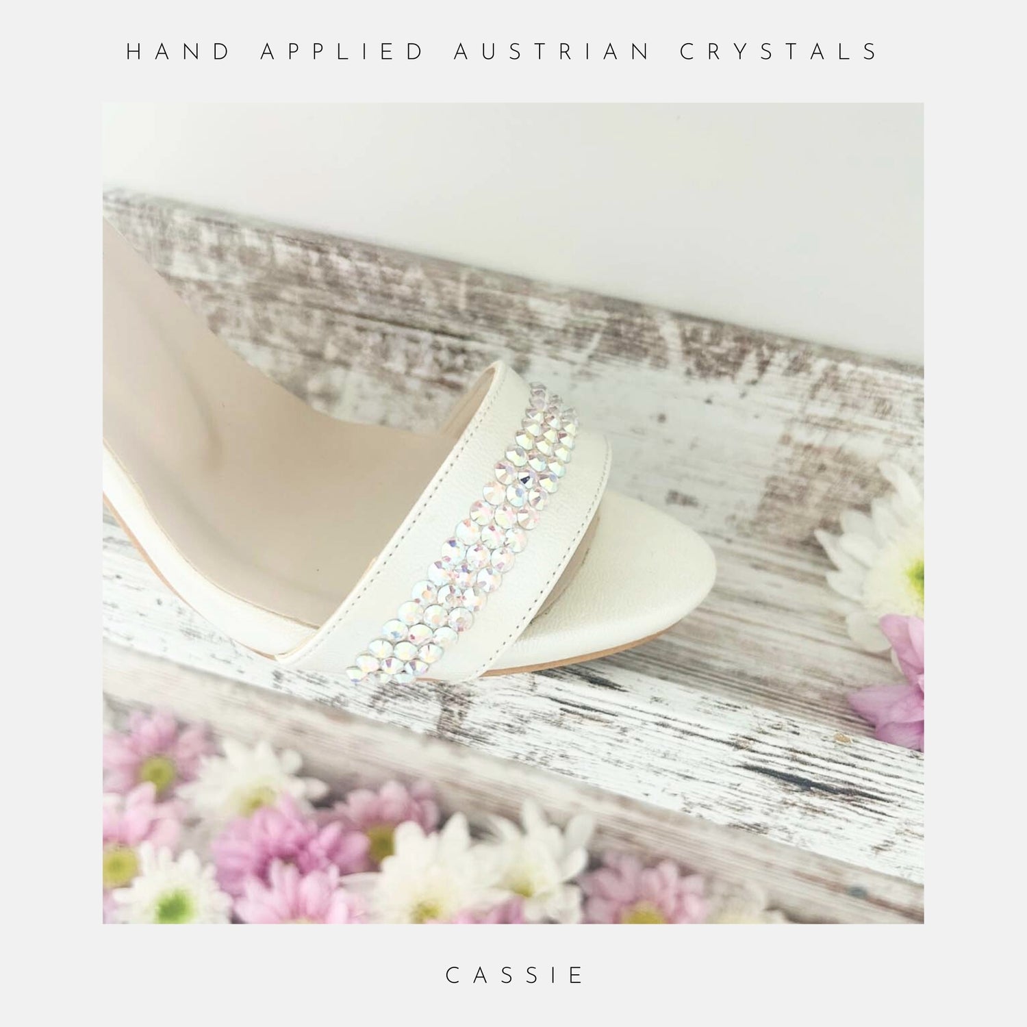 Ivory high-heeled shoes with hand-applied Austrian crystals on a wooden surface with flowers.