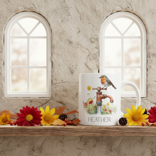 White mug with a design of a robin and flowers on a wooden surface with flowers and windows in the background.