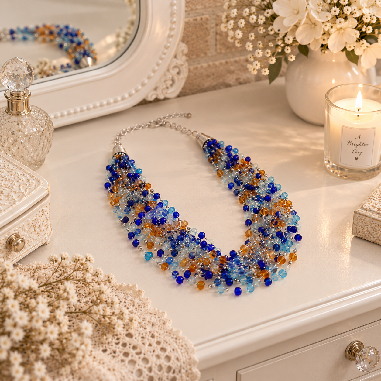 A multi-strand statement necklace with blue, orange, and clear faceted beads on a silver chain, styled on a white vanity table with a mirror, candle, and dried flowers.
