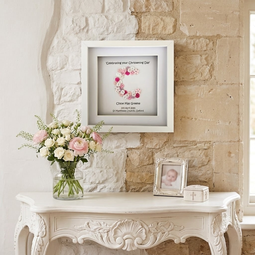 A white square framed print with a pink floral monogram and personalised text hangs on a light stone wall above a white ornate console table. A vase of pink and white flowers sits on the table, along with a small framed photo and a small box.