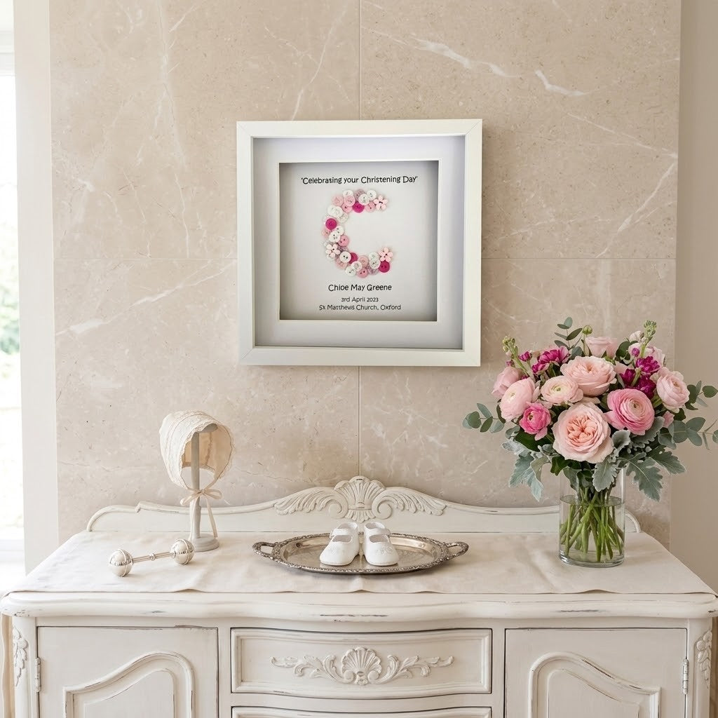A framed Christening Day print with a floral wreath design and personalised text, displayed on a beige marble wall above a white console table with flowers and baby shoes.