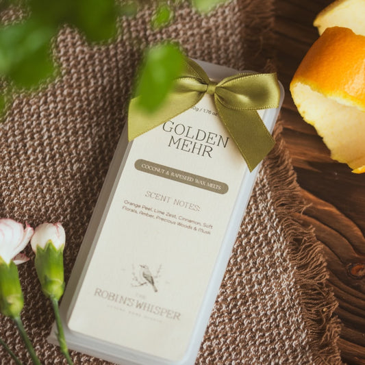 A boxed wax melt product labelled 'GOLDEN MEHR' by Robins Whisper, wrapped in a cream sleeve with a satin olive-gold ribbon bow, photographed on a woven brown textile with orange peel and flowers as styled props.
