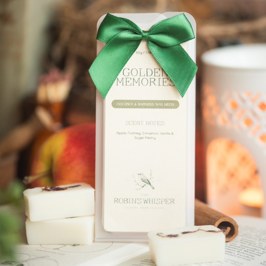 A boxed scented wax melt product labeled Golden Memories by Robins Whisper, tied with a green ribbon, standing upright with white wax bars placed in front on a tabletop. The scene is styled with warm blurred background elements, including a lantern and fruit.