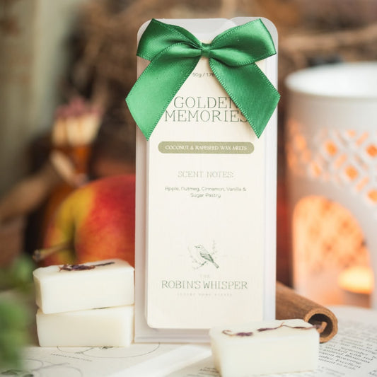 A boxed scented wax melt product labeled Golden Memories by Robins Whisper, tied with a green ribbon, standing upright with white wax bars placed in front on a tabletop. The scene is styled with warm blurred background elements, including a lantern and fruit.
