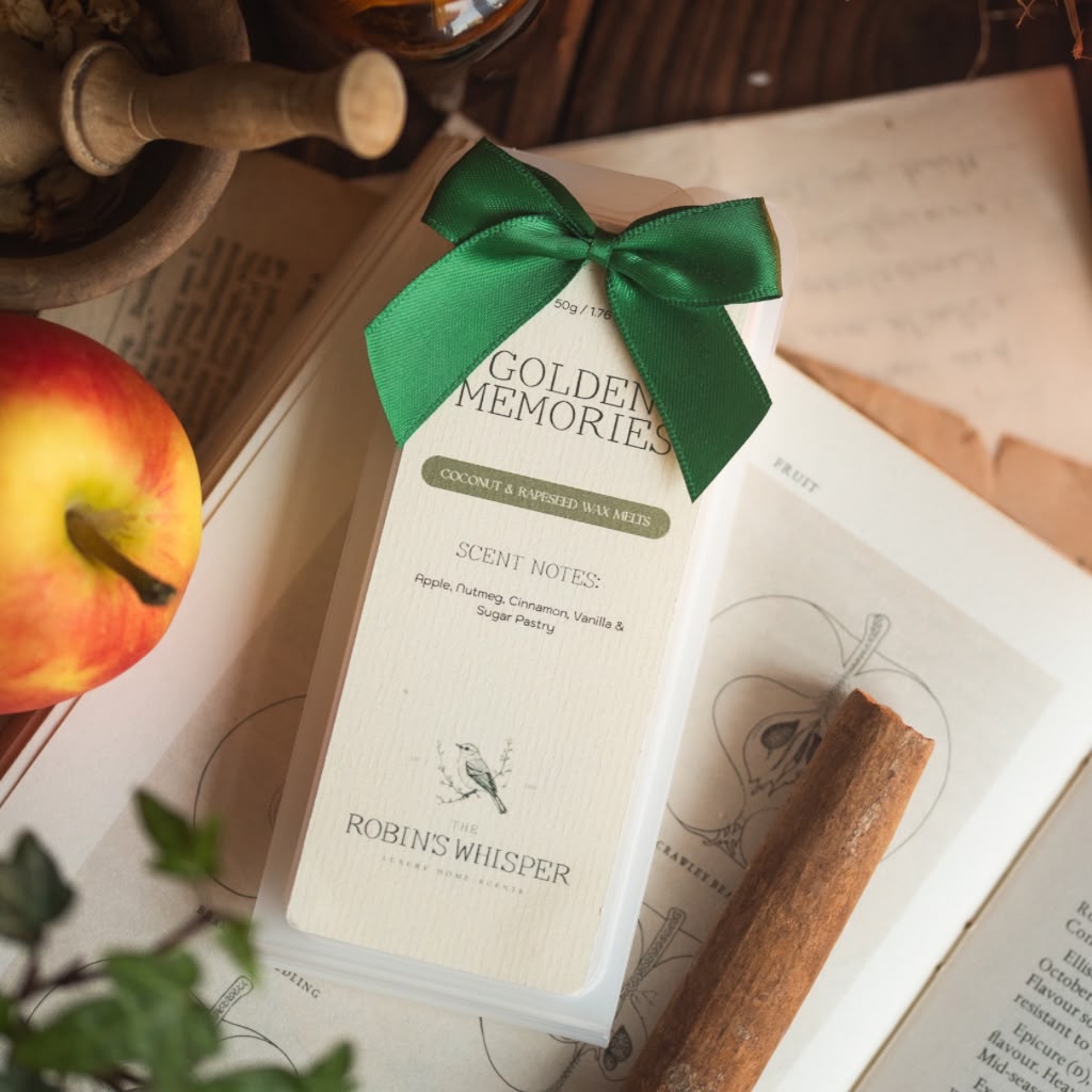A boxed wax melts product labelled 'Golden Memories' by Robins Whisper, wrapped in a white box with a green ribbon bow, styled on an open book with an apple, cinnamon stick, and botanical props nearby.