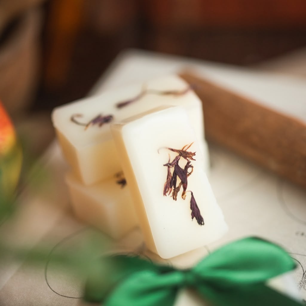 Three rectangular bars of wax melts stacked together, featuring dried botanical inclusions, presented on a light surface with a green ribbon bow in the foreground.
