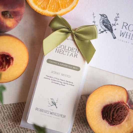 A packaged wax melt bar labelled 'Golden Nectar' by Robin's Whisper, tied with an olive green ribbon, styled with peach halves and an orange slice on a light surface.