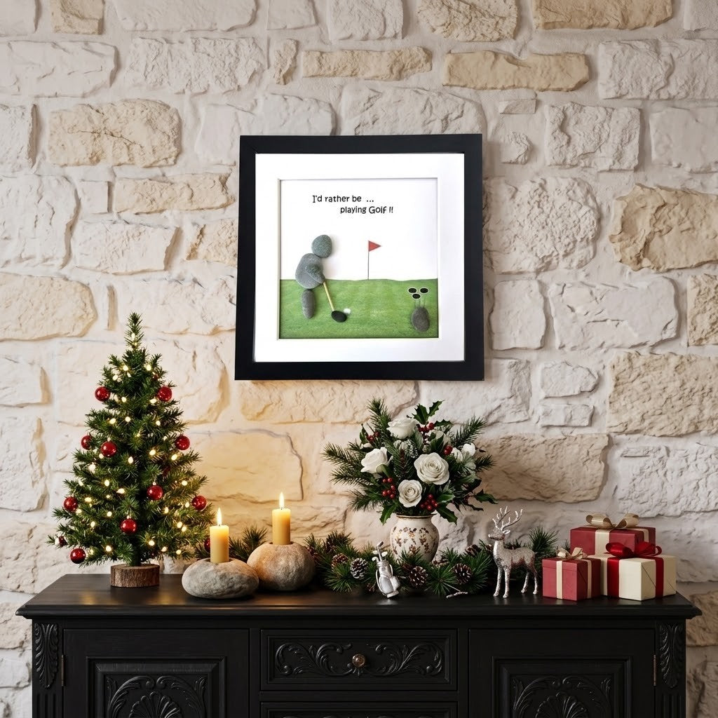 A framed art print featuring a whimsical golf illustration with the text 'I'd rather be playing Golf it' hangs on a light stone brick wall above a dark wooden console table decorated for Christmas with a small lit tree, candles, flowers, and wrapped gifts.