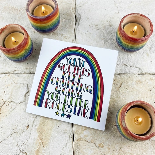 A square white greeting card with a colourful rainbow graphic and the text 'YOU'VE GOT THIS CHUFFING GOING YOU UTTER ROCKSTAR' lies on a light stone surface, surrounded by four lit candles in small rainbow-striped holders.