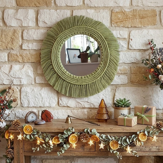 An olive green woven macrame round mirror with a thick fringe border hangs on a light stone brick wall above a rustic wooden mantel decorated with dried orange slices, pinecones, small plants, wrapped gifts, and warm string lights.