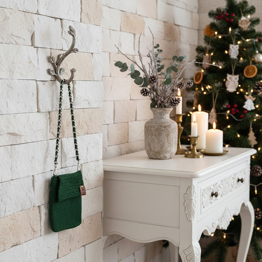 Decorative setting with a green bag hanging on a wall hook, a table with candles and a vase, and a Christmas tree in the background.