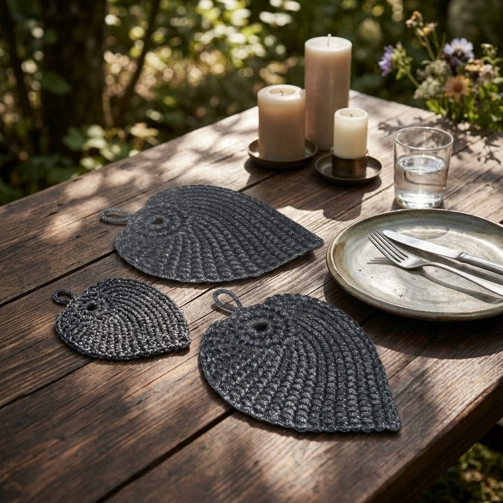 Three dark charcoal woven leaf-shaped placemats arranged on a rustic wooden dining table outdoors, with candles, a glass of water, and a plate with cutlery nearby.