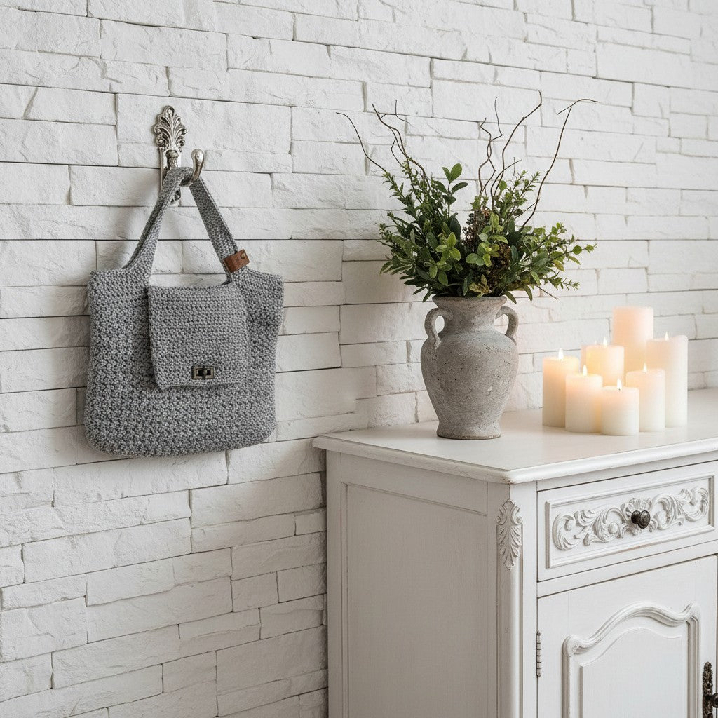 Grey crocheted bag hanging on a wall hook next to a white cabinet with candles and a vase.