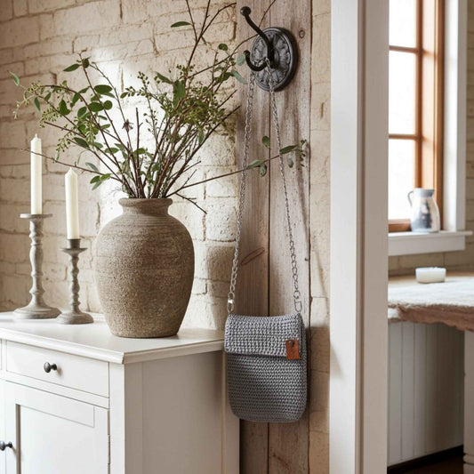 Grey woven bag hanging on a hook next to a vase with plants on a white cabinet.
