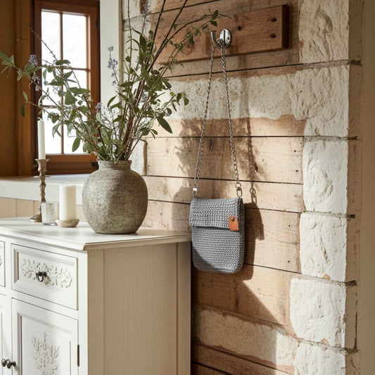 Grey woven handbag hanging on a wooden wall next to a vase with plants.