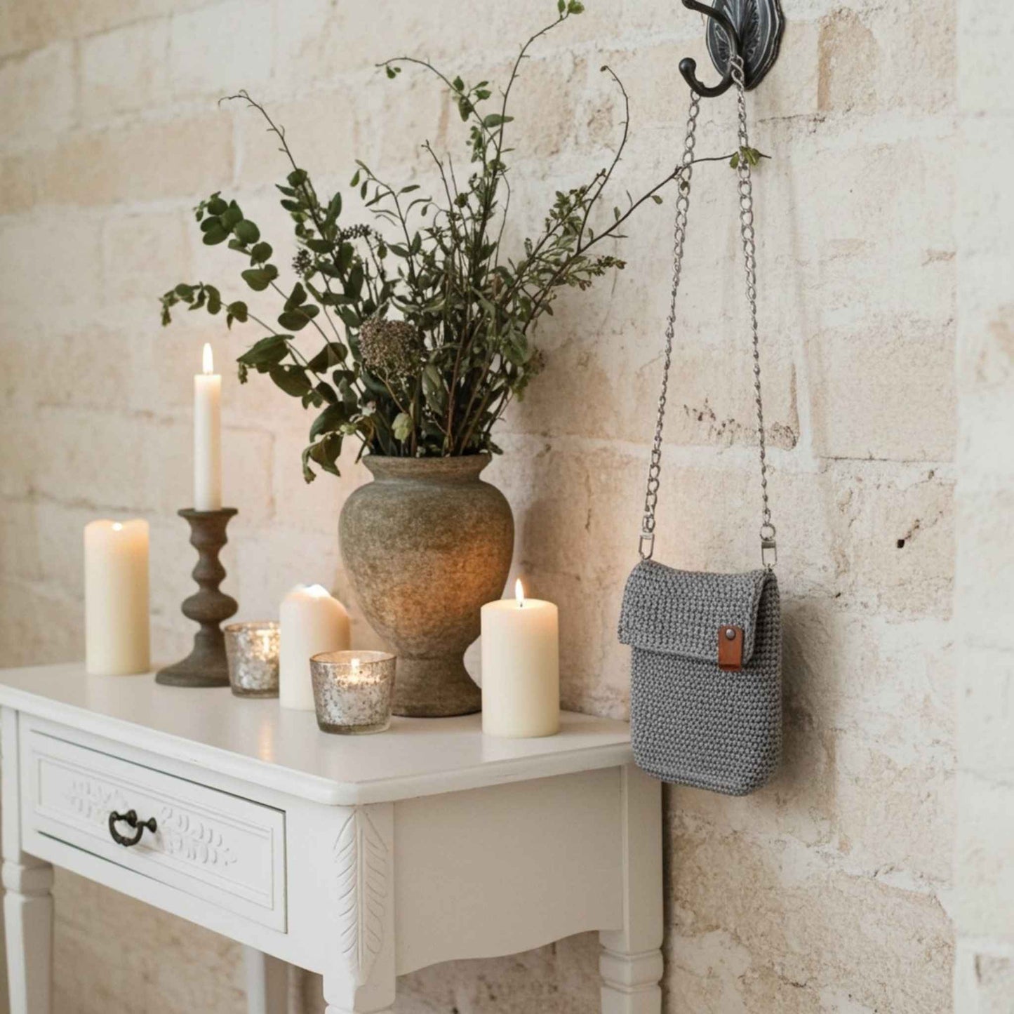 Grey knitted bag hanging on a wall next to a white console table with candles and a vase.