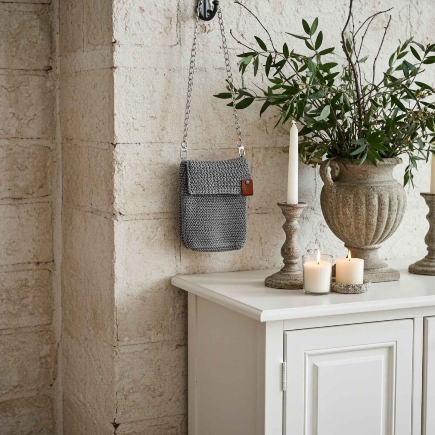 Grey woven bag hanging on a stone wall next to a white cabinet with candles and a plant.