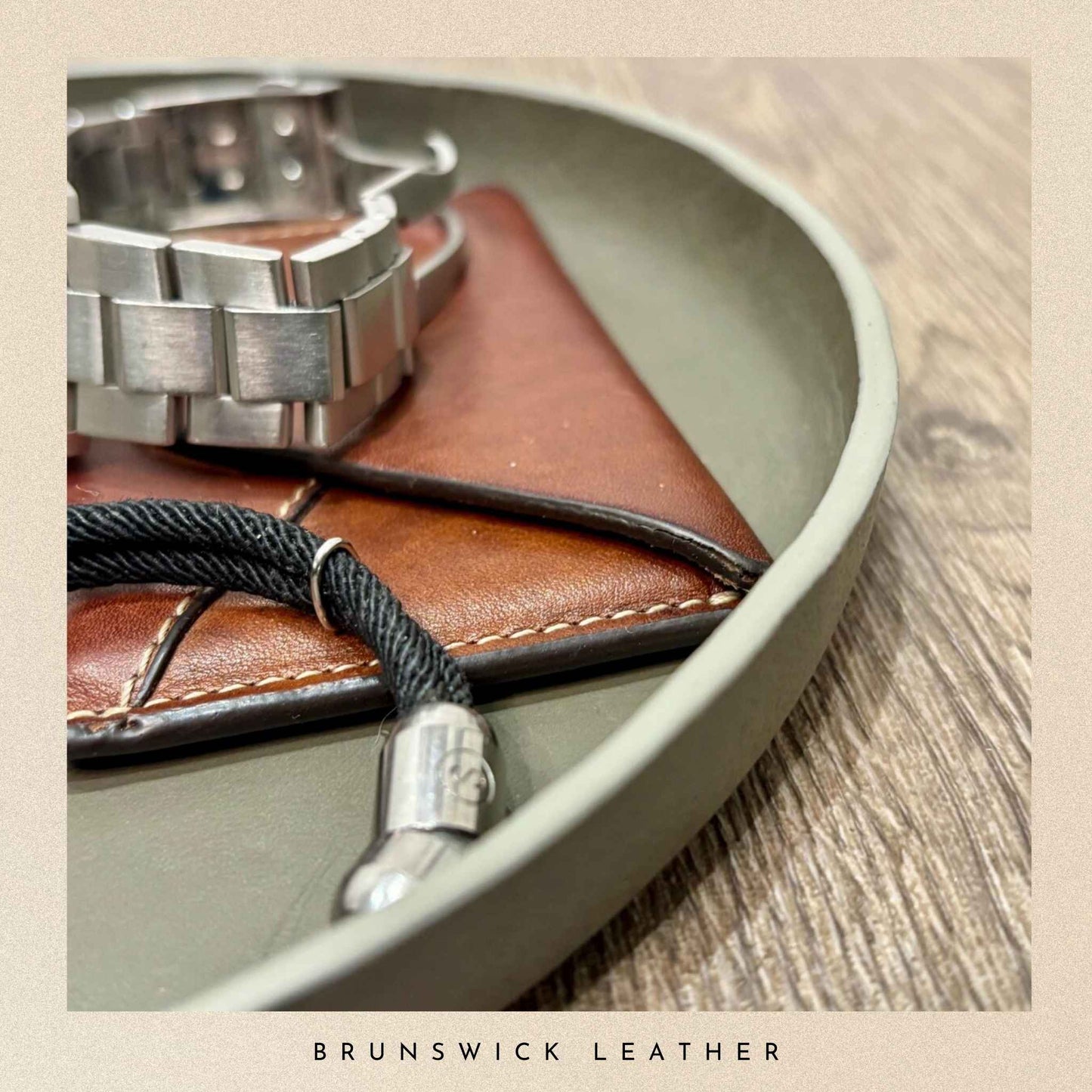 Brown leather wallet with silver watch and black lanyard on a grey leather valet tray, branded 'Brunswick Leather'.