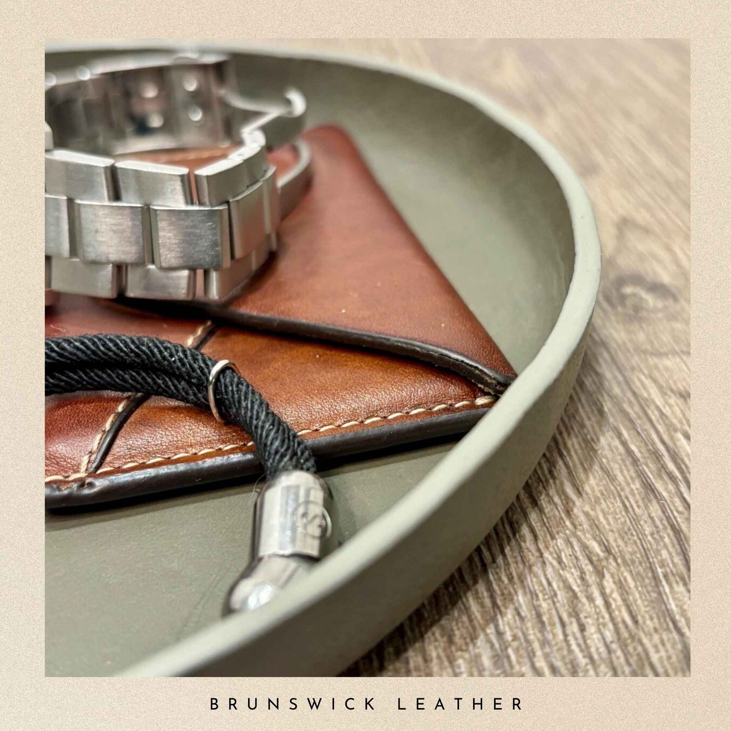 Brown leather wallet with silver watch and black lanyard on a grey leather valet tray, branded 'Brunswick Leather'.
