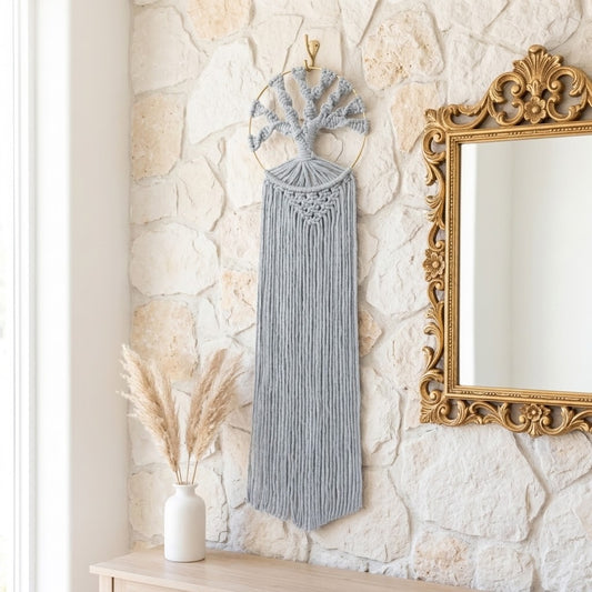 A grey macrame wall hanging with a tree design and long fringe, displayed on a rustic stone wall next to a gold-framed mirror and a small vase with dried grass.