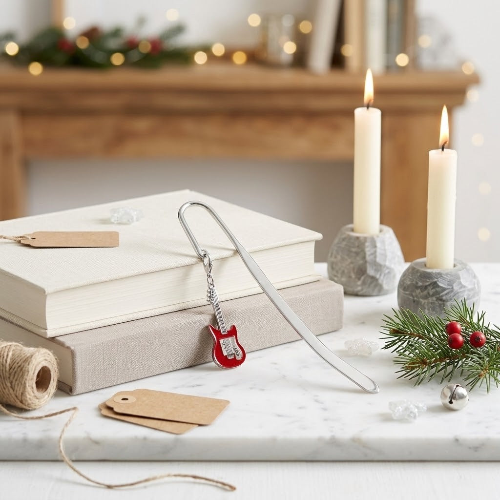 A silver metal bookmark shaped like a small red guitar rests on stacked books on a white marble surface, with candles, pine branches, and holiday decor in a softly blurred indoor setting behind it.