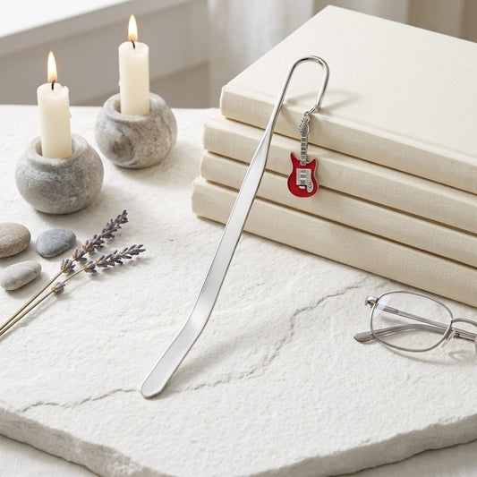 A silver metal bookmark shaped like a small red electric guitar is draped over a stack of cream-coloured books on a light stone surface, with candles, pebbles, lavender sprigs, and eyeglasses nearby.