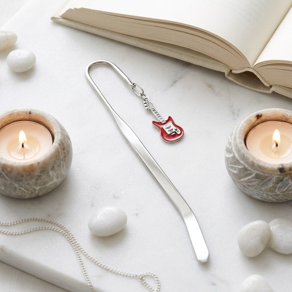 A silver metal bookmark shaped like a guitar headstock with a red and white pick detail, displayed on a white marble surface with lit candles, white stones, and an open book.