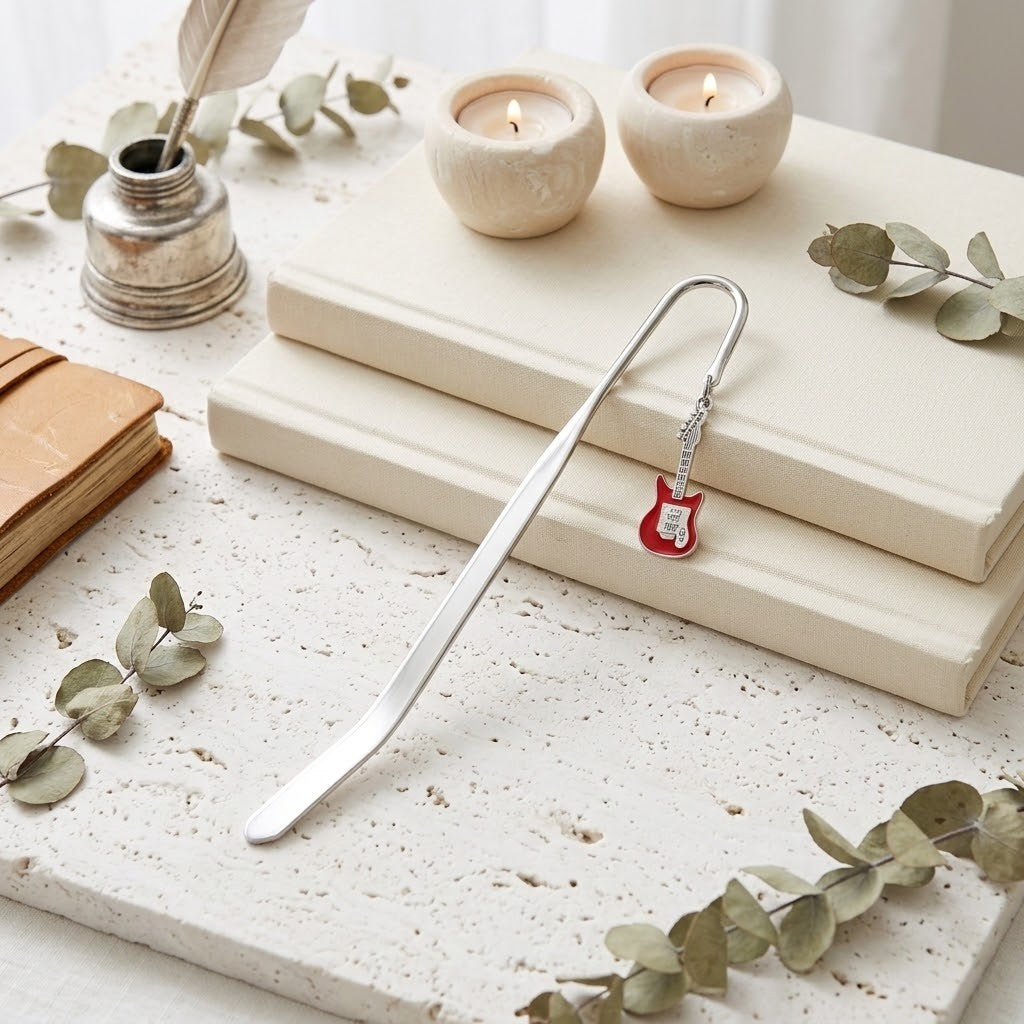 A silver metal bookmark with a small red guitar charm lies on stacked cream-coloured books, styled with eucalyptus leaves, candles, and a vintage ink bottle.
