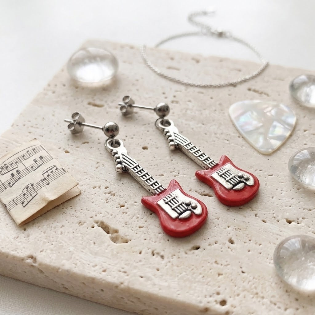 Red and silver guitar-shaped drop earrings displayed on a textured beige stone slab with decorative props including a small sheet of music, a guitar pick, and clear beads.