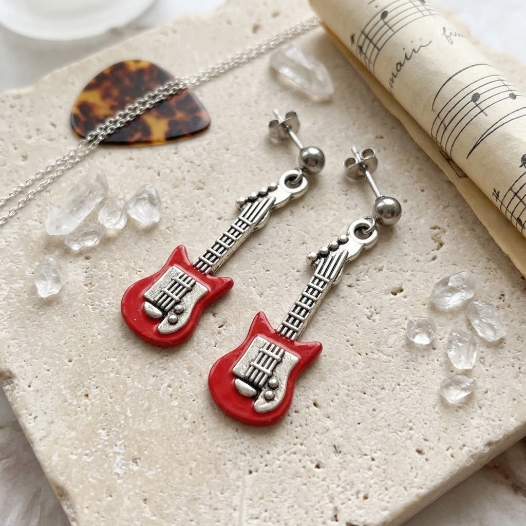 Red electric guitar stud earrings with silver-tone necks and heads, displayed on a textured stone surface with a guitar pick, chain, sheet music, and clear crystals.