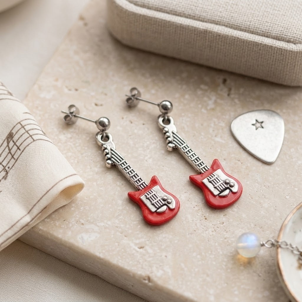A pair of dangling earrings shaped like red electric guitars with silver-tone necks and posts, displayed on a beige stone slab with fabric and a guitar-pick-shaped accessory nearby.