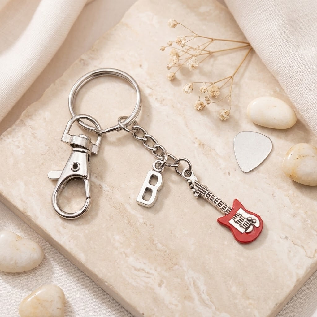A silver metal keychain with a lobster clasp, a hanging letter 'B' charm, and a guitar-shaped charm, displayed on a beige stone tile with decorative stones and dried flowers.