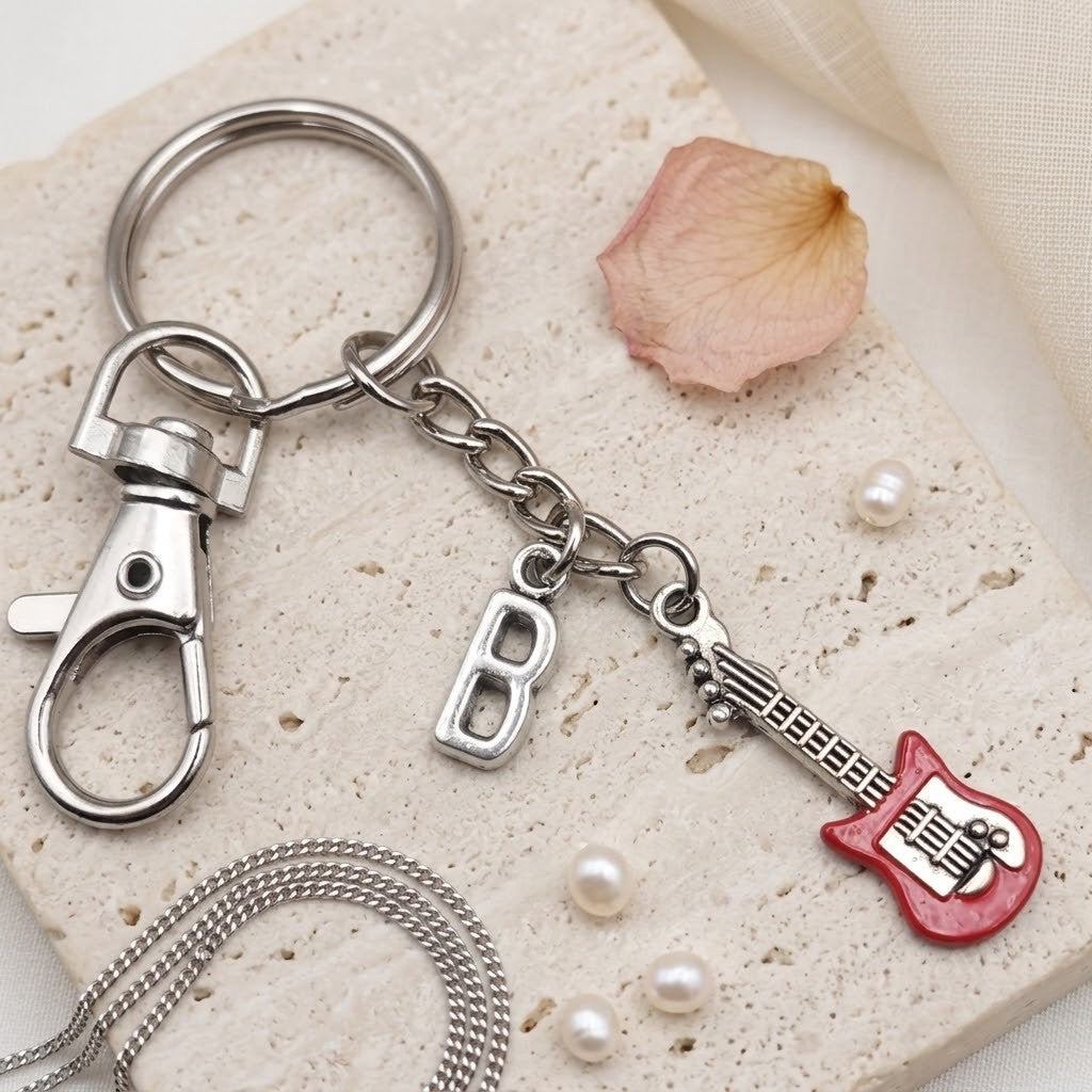 A silver-tone metal keychain with a lobster clasp, a letter 'B' charm, and a red guitar-shaped charm, styled on a textured beige stone tile with pearl beads and a dried flower petal.