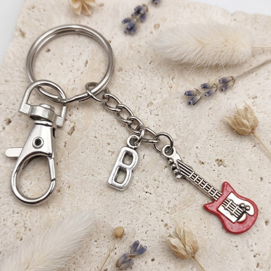 A silver metal keychain featuring a lobster clasp, a letter 'B' charm, and a guitar-shaped charm with a red body, displayed on a textured beige stone surface with dried flowers.