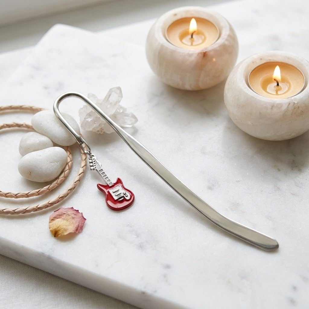 A silver metal bookmark shaped like a guitar pick with a small red guitar charm, styled on a white marble tray with white stones, a braided cord, a crystal, and two lit candle holders.